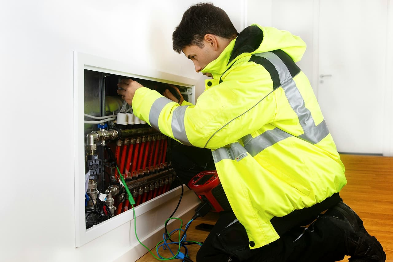 A technician in a high-visibility jacket kneels at an open in-wall heating manifold in a finished apartment, testing it with a meter.