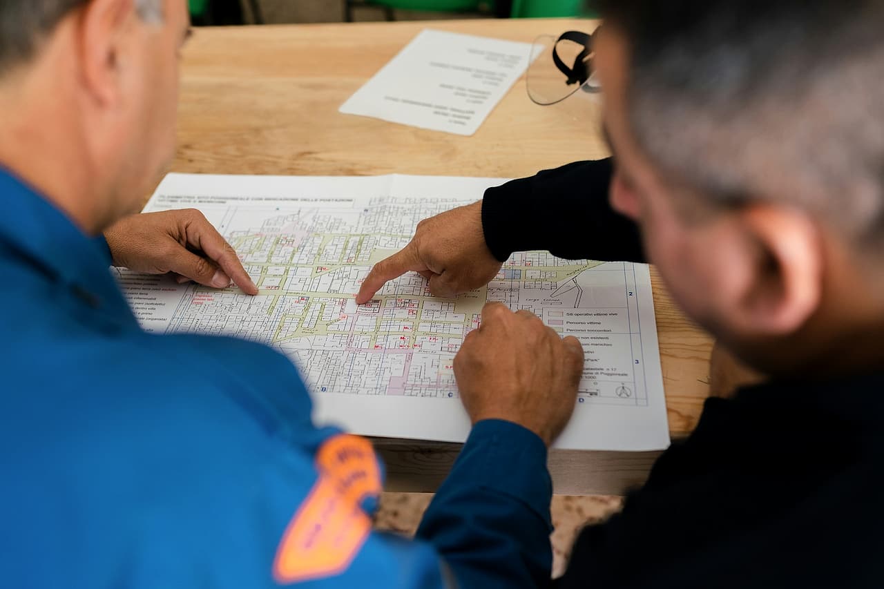 Two people seen from behind point to places on a large printed plan spread across a meeting table.