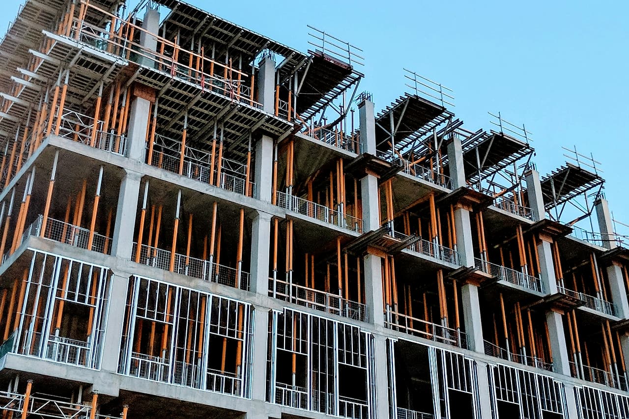 A multi-storey concrete frame under construction, with steel-stud exterior walls going in and shoring still in place, under a blue sky.