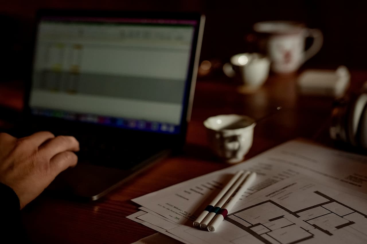 A hand on a laptop showing a spreadsheet, beside printed floor plans, marker pens and coffee cups on a desk in warm light.