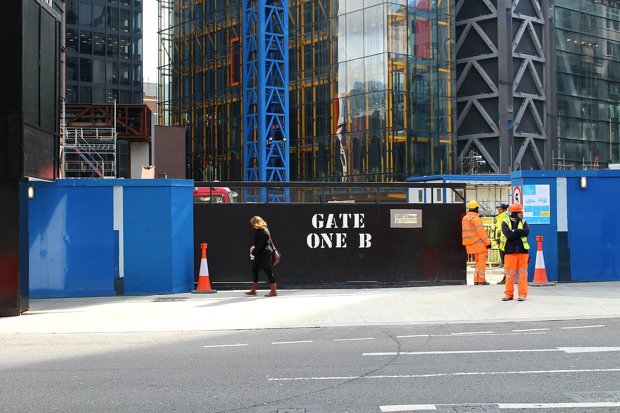 Three construction workers in hard hats and high-visibility clothing wait at a closed site gate marked Gate One B on a city street.
