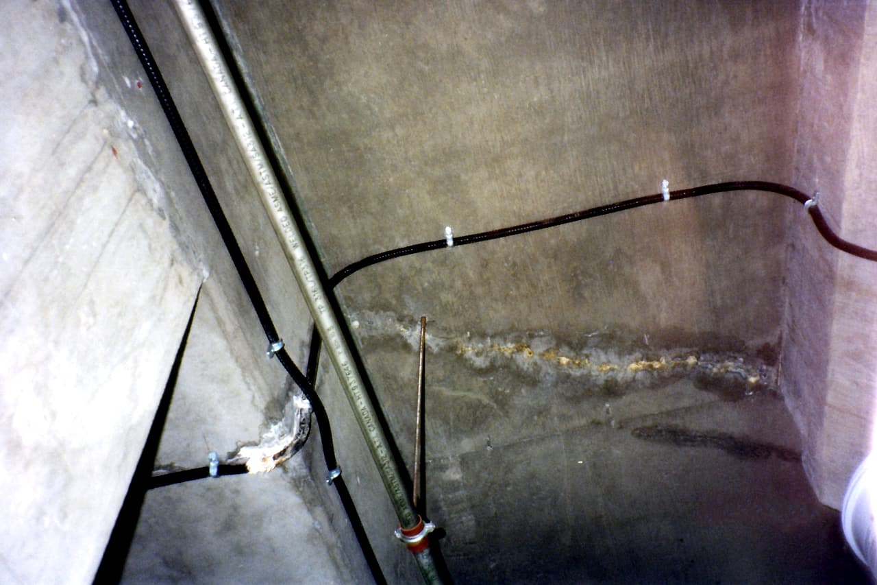 White mineral deposits and darker stains cross the underside of a concrete slab beside fixed pipes and conduits.