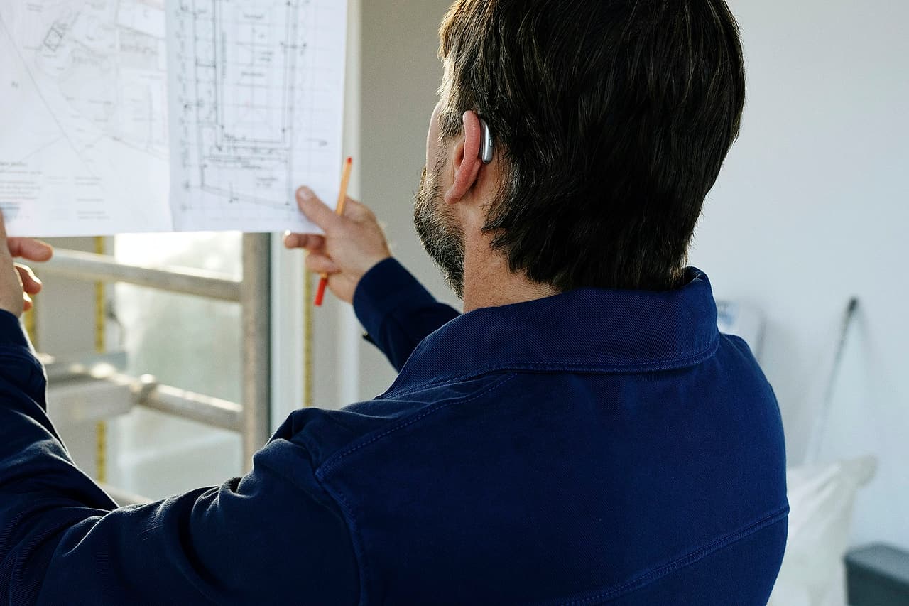 A person seen from behind points to a construction drawing while another person gestures beside the sheet.