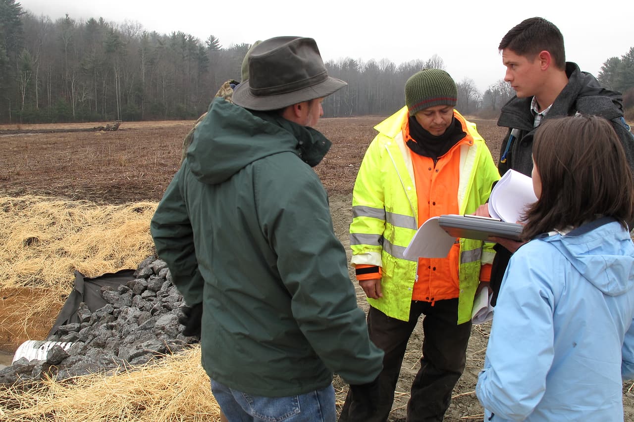 Four people review construction plans together beside a rural work area.