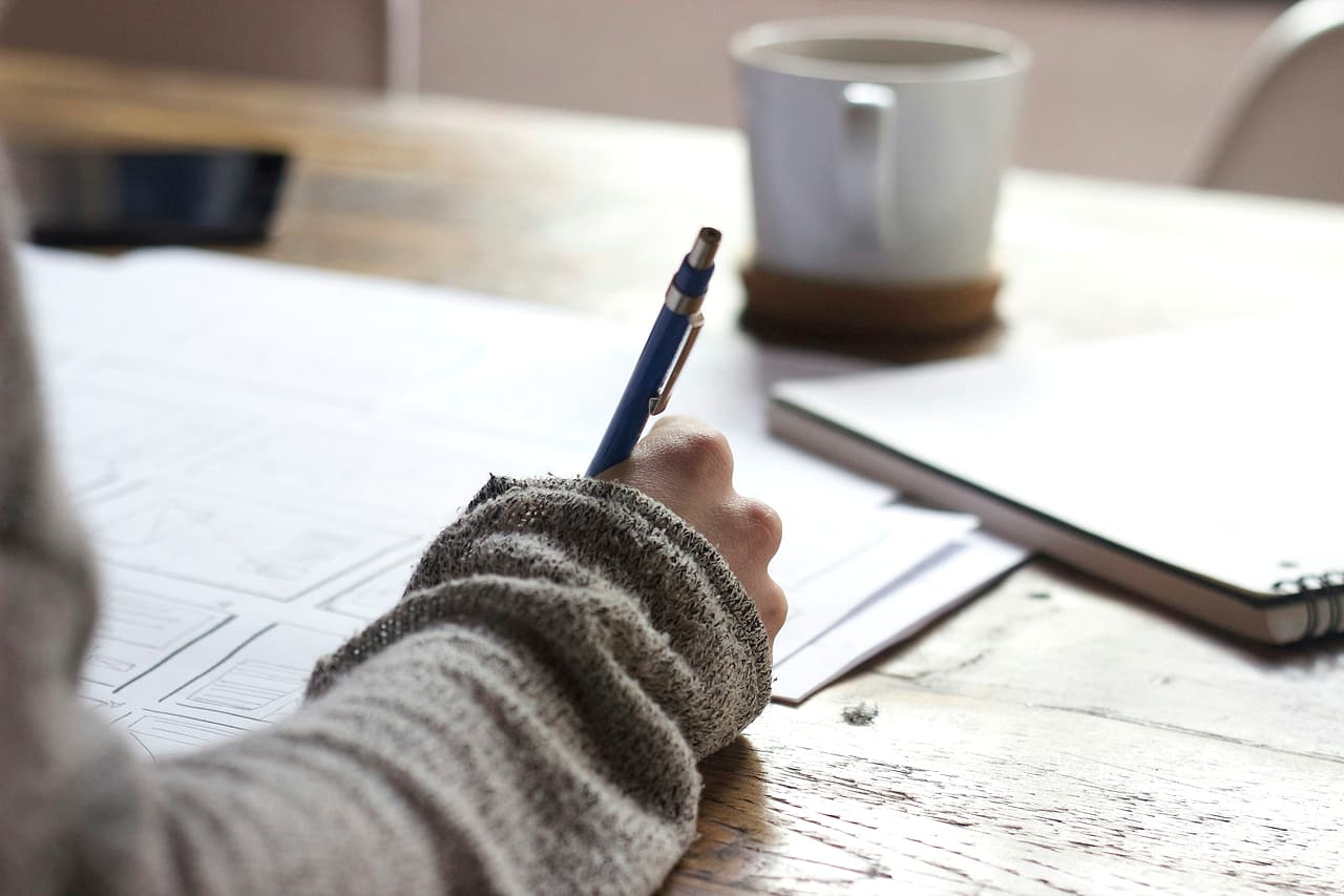 A hand writes on paper at a wooden table beside a notebook and cup.