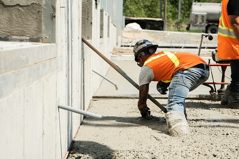 A concrete finisher in an orange vest, hard hat and gloves screeds a freshly poured slab beside a wall footing, a mixer truck behind.