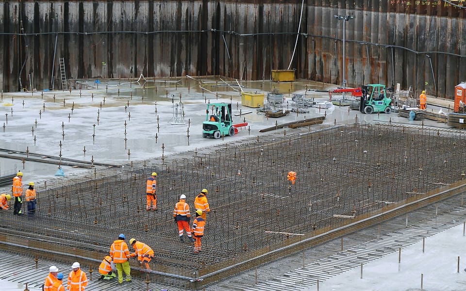 A crew in orange hi-vis clothing lays the reinforcement of a raft slab at the bottom of a sheet-piled excavation, two forklifts parked on the blinding.