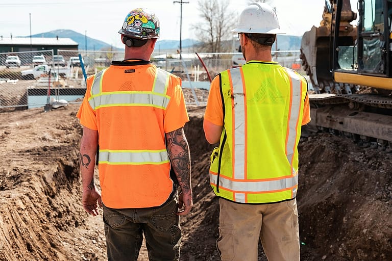 Two workers in hi-vis vests and hard hats, seen from behind, look over an open excavation, an excavator parked on the far side.