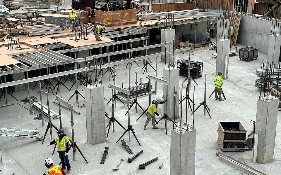 A formwork and shoring deck seen from above: concrete columns, steel shores and a crew in hi-vis vests setting the next pour.