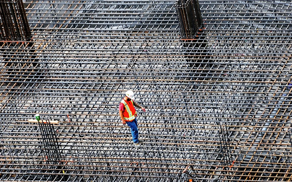 Seen from above, a lone worker in a white hard hat and orange vest walks across a reinforcement mat, checking it before the pour.