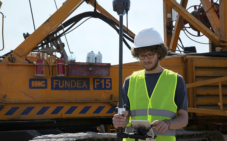 A surveyor in a white hard hat and hi-vis vest reads a tablet while holding a GNSS rover, a piling rig behind him.